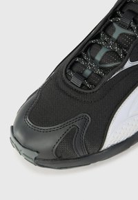 Zapato deportivo negro con parte superior de tela texturizada, superposiciones sintéticas, acentos reflectantes y costuras distintivas en la puntera.