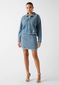 Ensemble veste en jean bleu clair et jupe, avec bords effilochés, boutons dorés et poches, complété par des sandales argentées à lanières.