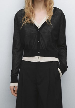 Person wearing layered black cardigan over beige shirt with black wide-leg pants, one hand in pocket, against plain light background.