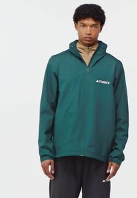 Young man wearing a teal Terrex zip-up jacket over a beige top and black pants, standing against a plain white background.