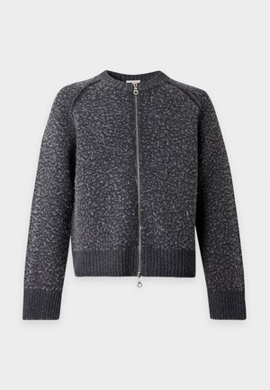 Homme portant un pull zippé gris foncé texturé, une chemise blanche boutonnée, un pantalon ample gris foncé et des chaussures bordeaux avec des lacets rouges, debout devant un fond blanc.