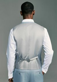 Gilet en satin gris clair avec une texture lisse, détail de boucle au dos et coupe ajustée, associé à une chemise blanche à manches longues.