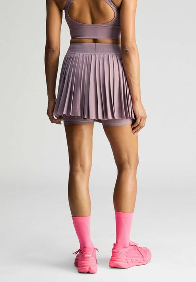Woman standing showing back view wearing mauve pleated skirt with built-in shorts, matching sports bra, bright pink socks, and sneakers.
