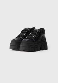 Ténis de plataforma pretos feitos de couro liso, com ilhós metálicos, solas de borracha grossas e uma marcação em relevo na parte frontal.