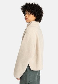 Joven con cabello rizado que lleva una chaqueta de forro polar beige claro y pantalones gris oscuro, mirándose por encima del hombro.