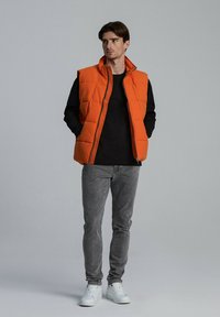 Pomarańczowa pikowana kamizelka puchowa z wysokim kołnierzem, noszona na czarnej koszulce z długim rękawem. Szare dżinsy i białe trampki dopełniają strój.