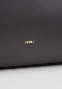 Musta nahkapinta, jossa on pieni kultainen Furla-logo keskellä, valkoisen taustan alareunassa.