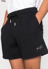 Shorts en coton noir avec une ceinture élastique, cordons de serrage roses et poches latérales. Présente un texte brodé rose sur l'ourlet.