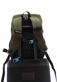 Mochila verde con respaldo de malla negra, correas ajustables y herrajes azules. Tiene un asa en la parte superior y está montada sobre una maleta negra.