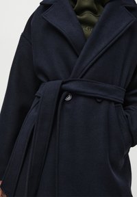 Palton din amestec de lână de culoare bleumarin, cu design înfășurat, rever lat, curea și nasturi mari. Textură netedă și croială lejeră pentru confort.