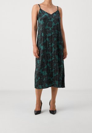 Frau trägt ein grünes, plissiertes Midikleid mit schwarzem Blumenmuster und dünnen Trägern, kombiniert mit schwarzen spitzen High Heels.