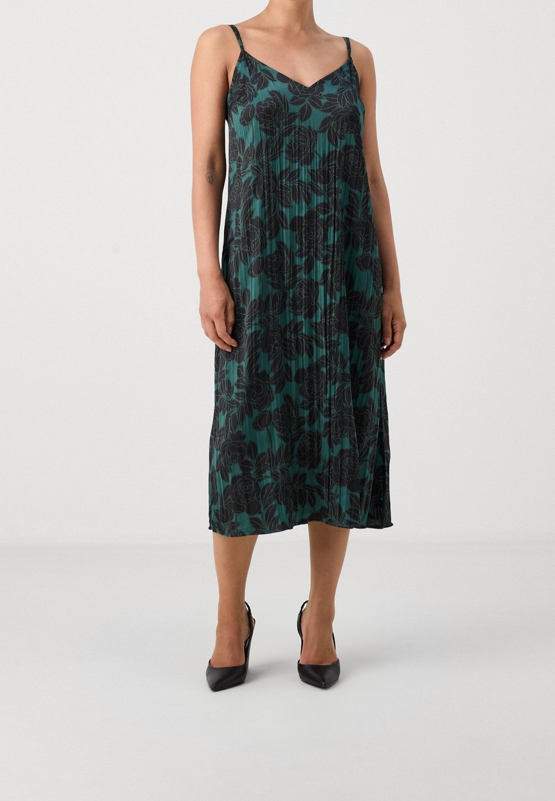 Femme portant une robe midi plissée verte avec imprimé floral noir et fines bretelles, associée à des talons noirs pointus.