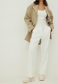 Bežový vlněný kabát se širokými klopy, bílý top na ramínka, přizpůsobené bílé kalhoty a robustní b�éžové boty. Texturovaná tkanina a čisté linie.