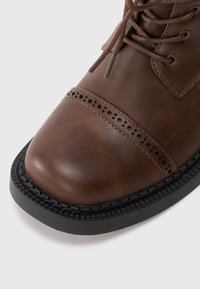 Stivale in pelle marrone con punta tondeggiante, superficie texturizzata, dettagli perforati e suola in gomma nera. Dettagli dei lacci visibili sulla parte superiore.