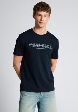 Junger Mann mit lockigem Haar, der ein schwarzes T-Shirt mit dem Schriftzug "ORIGINAL" und hellblaue Jeans trägt, steht vor einem schlichten, hellen Hintergrund.