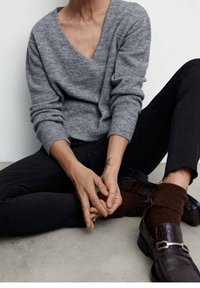 Suéter gris de cuello en V hecho de material suave, combinado con jeans negros ajustados. Un calcetín marrón con relieve y unos zapatos de loafer oscuros con detalle de hebilla son visibles.