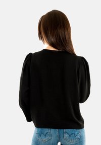 Femme avec de longs cheveux bruns portant un haut noir à manches longues et un jean bleu, vue de dos sur un fond blanc uni.