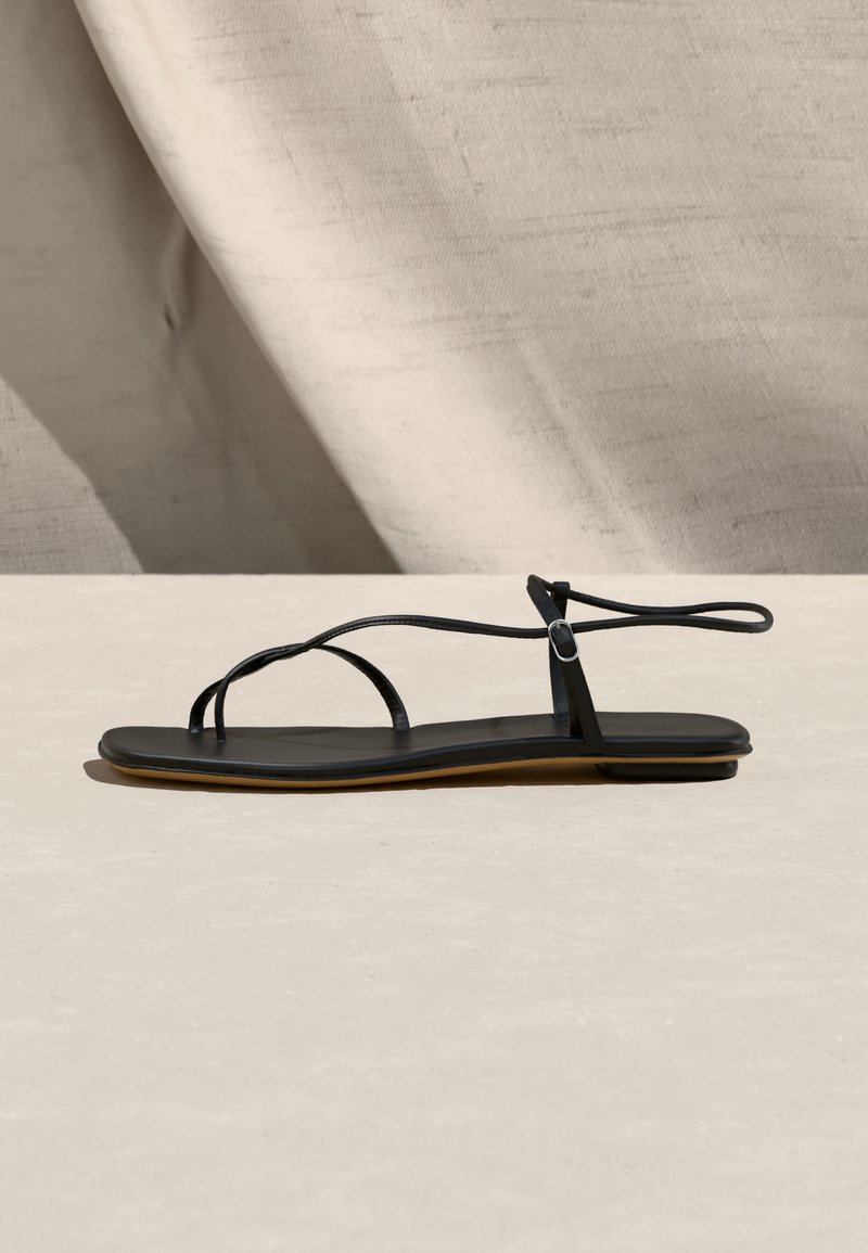 Μαύρο φλατ σανδάλι με λεπτούς ιμάντες και αγκράφα στον αστράγαλο, τοποθετημένο σε μπεζ υφασμάτινο φόντο και επιφάνεια.