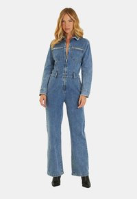 Tuta in denim con maniche lunghe, chiusura frontale con zip, due tasche sul petto e vita affusolata. Colore azzurro chiaro e design a gamba larga. Stivali neri alla caviglia.