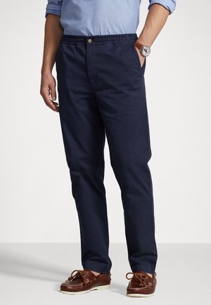 Homme portant un pantalon décontracté bleu marine avec taille élastique, une chemise bleu clair aux manches retroussées, une montre en cuir marron et des chaussures bateau marron.