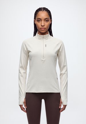 Une femme porte un haut de sport blanc à manches longues et à demi-zip, ainsi qu’un legging foncé, sur un fond clair uni.