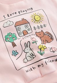 Sudadera rosa con ilustraciones divertidas: una casa, el sol, una nube, un conejo, un gato, flores y árboles, con el texto "Me encanta jugar con mi amigo."