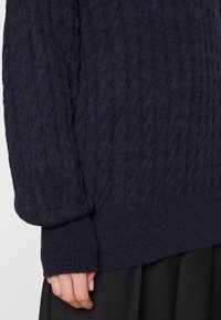 Pull en maille torsadée bleu marine avec une surface texturée, doté de poignets et d'un ourlet côtelés. Le tissu semble doux et épais, offrant une meilleure chaleur.