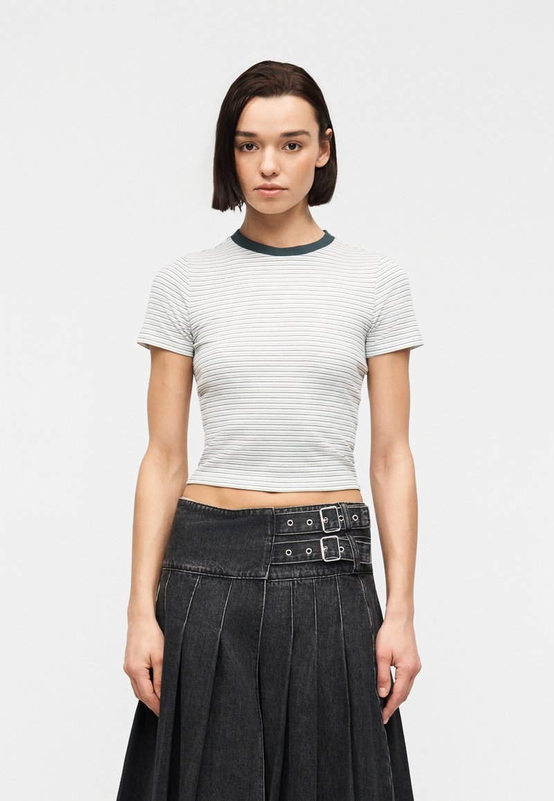 Jeune femme aux cheveux courts bruns portant un T-shirt blanc ajusté à rayures et une jupe noire plissée taille haute avec des détails de boucle.