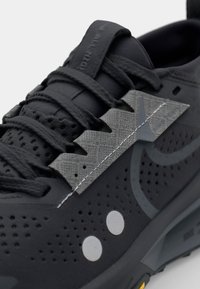 Black athletic shoe with perforated upper, textured grey accents, and reflective details. Features grey laces and a cushioned sole.