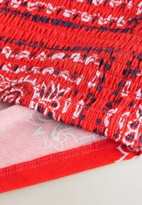 Sarkanā auduma tuvplāns ar saplēstu faktūru un baltām un tumši zila abstraktām rakstām, redzama kārtīga šuve un daļa no zem tās esošās gaiši rozā rakstainās oderes.