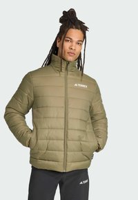 Ung mand med dreadlocks iført en fyldig olivengrøn Terrex jakke, står mod en lys baggrund, hænderne i lommerne, og kigger selvsikkert ind i kameraet.