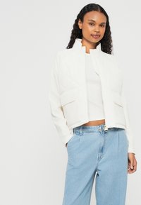 Casaco branco acolchoado com gola alta, duas bolsos frontais e um design curto, combinado com jeans de perna reta azul claro.