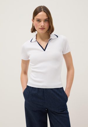 Mujer joven con cabello corto vistiendo una camisa polo blanca de manga corta con ribete azul marino y pantalones anchos azul oscuro, con las manos en los bolsillos, fondo neutro.