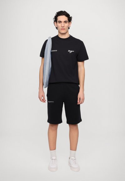 Zwarte katoenen t-shirt met tekst en ontwerp, gecombineerd met zwarte shorts, een denim jas over de schouder en witte sneakers.