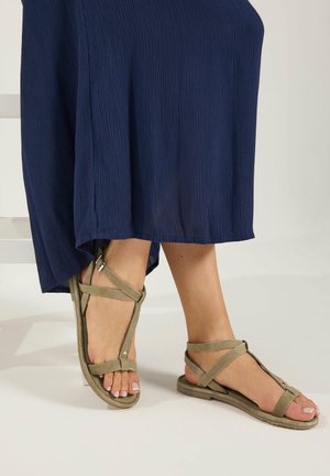 Füße, die beige flache Riemchensandalen tragen, kombiniert mit einem marineblauen, strukturierten langen Rock vor weißem Hintergrund.