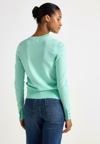 Mintgrøn striktrøje med ribbet kant og manchetter, har guldknapdetaljer på ærmerne, parret med mørkeblå denimjeans.