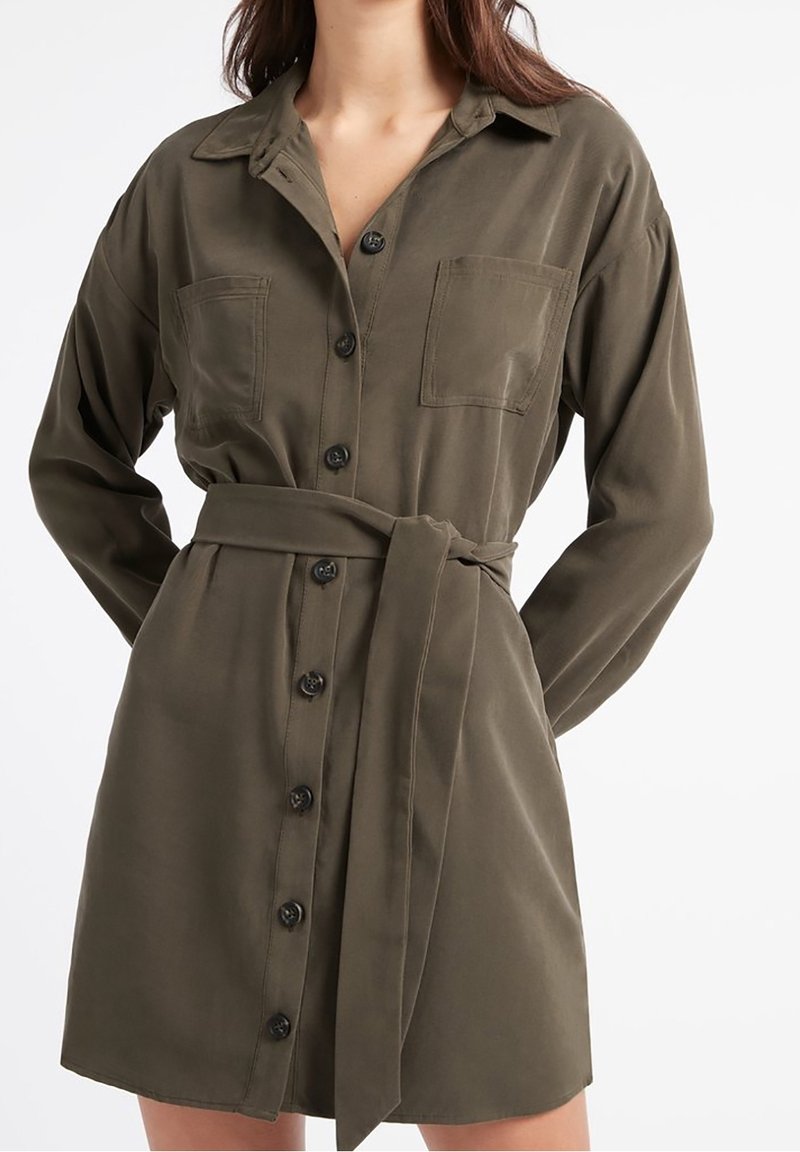 Vestido camisa verde oliva con cuello, frente abotonado, dos bolsillos en el pecho y un cinturón a juego atado en la cintura. Textura de tela suave.