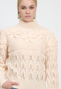 Maglione lavorato a maglia in morbido beige, con motivi floreali sulle spalle e un colletto a coste. Design a traforo nella parte inferiore.
