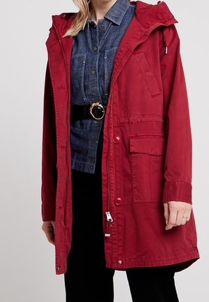 Frau trägt eine rote Kapuzenjacke über einem denimblauen Hemd mit Knöpfen, dazu einen schwarzen Gürtel und schwarze Hosen.