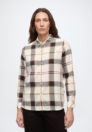 Femme aux longs cheveux ondulés portant une chemise à carreaux beige, marron, grise et moutarde boutonnée et un pantalon noir, debout devant un fond blanc.