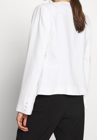 Femme portant un blazer blanc ajusté avec trois boutons sur la manche et un pantalon noir, vue de dos et de profil.
