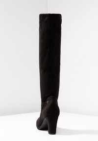 Botas de tacón alto de ante negro hasta la rodilla con una textura suave y una punta redondeada, que presentan un pequeño acento circular cerca del tacón.
