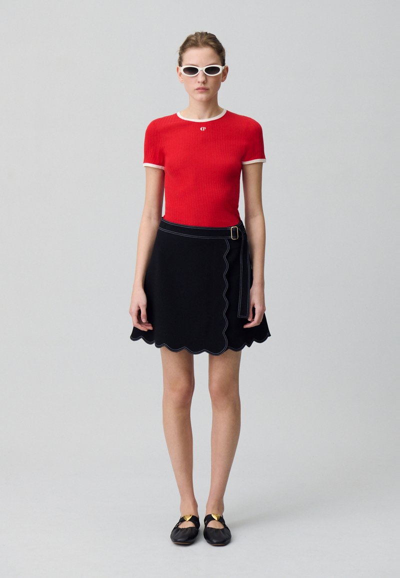 Haut court à manches courtes rayé rouge avec finitions blanches, associé à une jupe noire à ourlet festonné. Le mannequin porte des lunettes de soleil blanches et des chaussures noires avec des accents dorés.