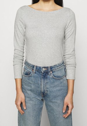 Person iført en tætsiddende lysegrå langærmet top, stoppet ned i højttaljede blå denimjeans med hænder afslappet langs siderne.