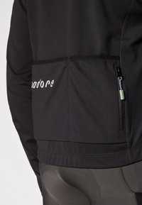 Čierny dlhý rukáv top s vreckom na zips na chrbte. Text "madore" v kontrastnej farbe blízko vrecka. Hladký, elastický materiál.