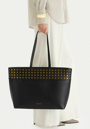 Osoba v bílých kalhotách a pruhovaném tričku drží černou koženou kabelku s kovovými zlatými cvoky a logem Badura.