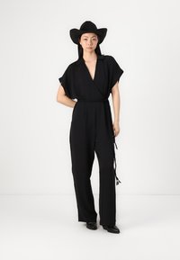 Svart, kortermet jumpsuit med V-hals, belted midje, vide ben og en glatt tekstur. Kombinert med en bredbrimmet svart hatt.