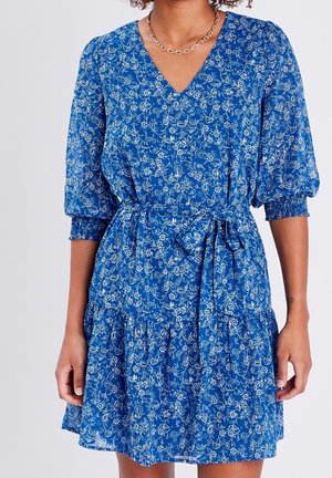 Femme portant une robe bleue à col en V avec motifs floraux, manches bouffantes, poignets élastiques et une ceinture assortie nouée à la taille.