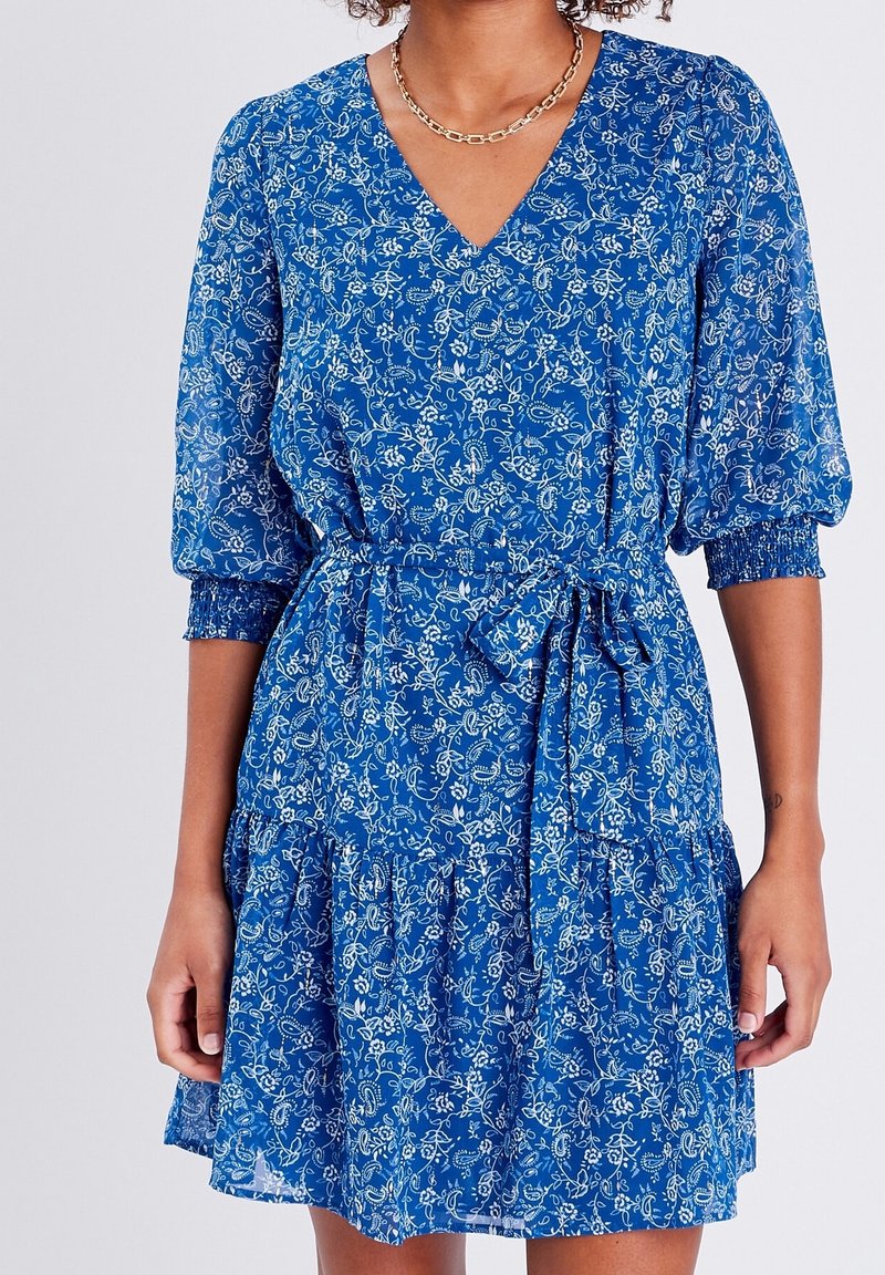 Femme portant une robe bleue à col en V avec motifs floraux, manches bouffantes, poignets élastiques et une ceinture assortie nouée à la taille.