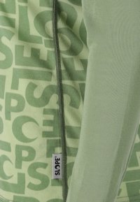 Groen langemouw top met een rits en grote, patroon tekst in een lichtere groen. Het label aan de zijkant toont de merknaam "SLOPE."